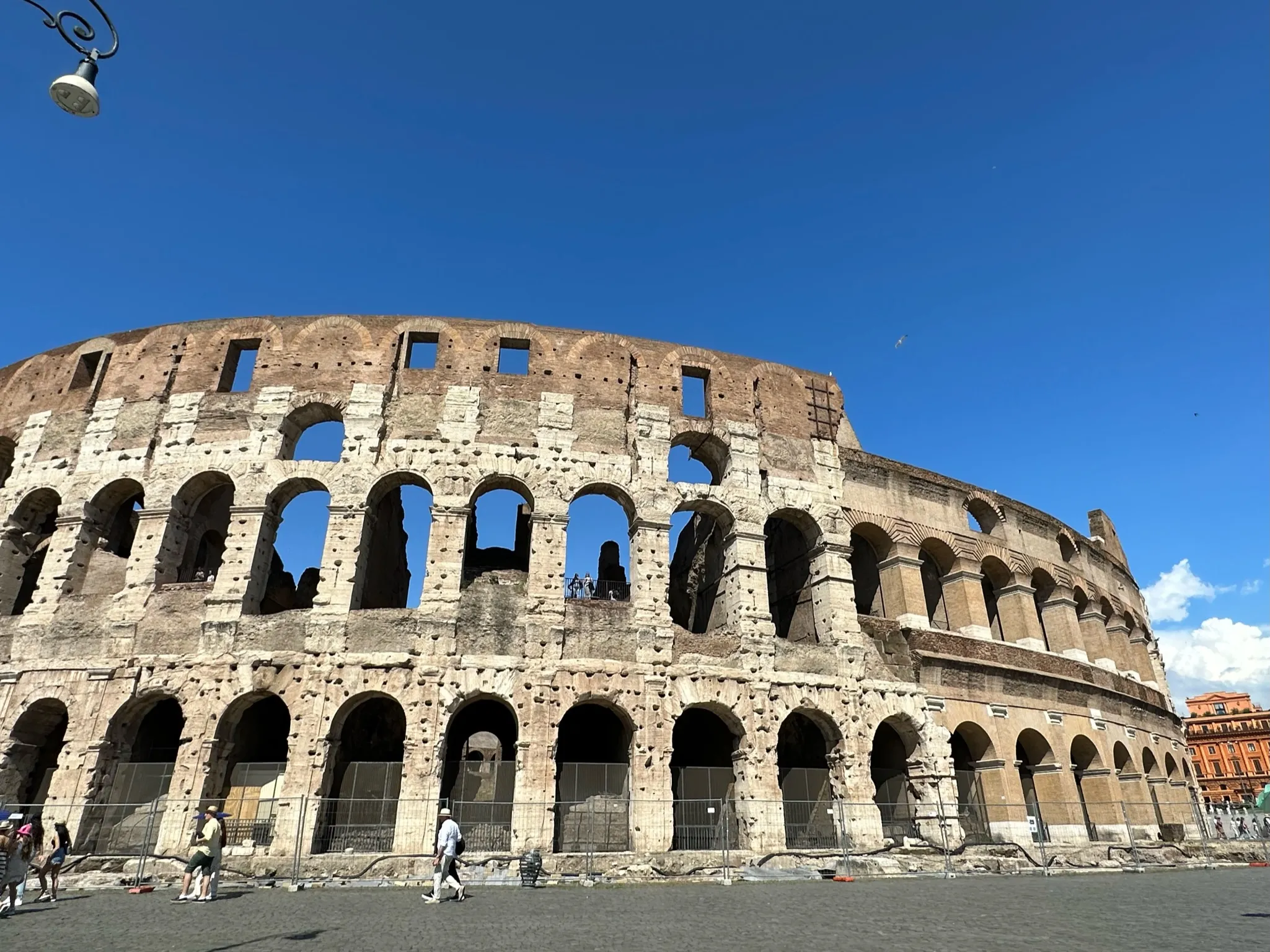 Wide view of the ancient Colosseum in Rome under a clear blue sky with few people walking nearby.