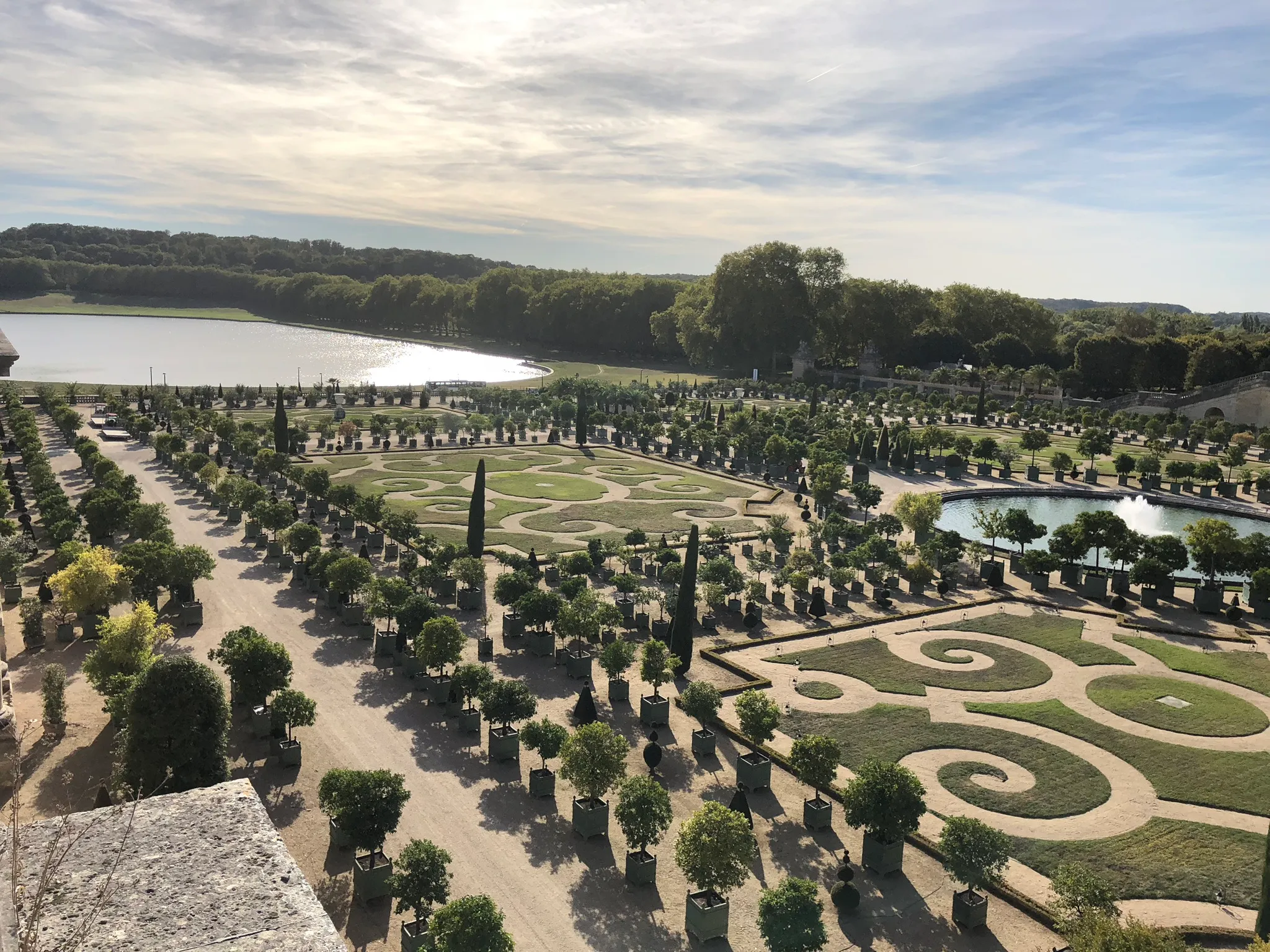 Aerial view of a formal garden with symmetrical patterned grass beds, numerous potted trees, a central fountain, and a large reflective pond surrounded by dense trees.