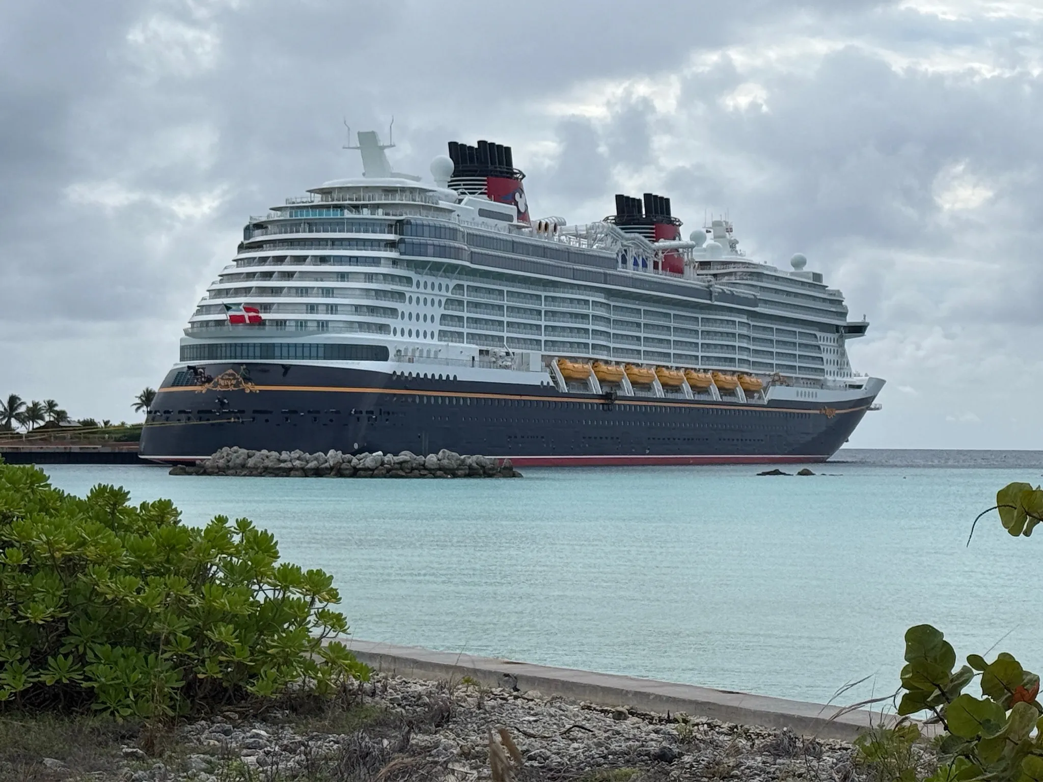 Large Disney cruise ship docked near shore under a cloudy sky, with clear turquoise water and tropical vegetation in foreground.