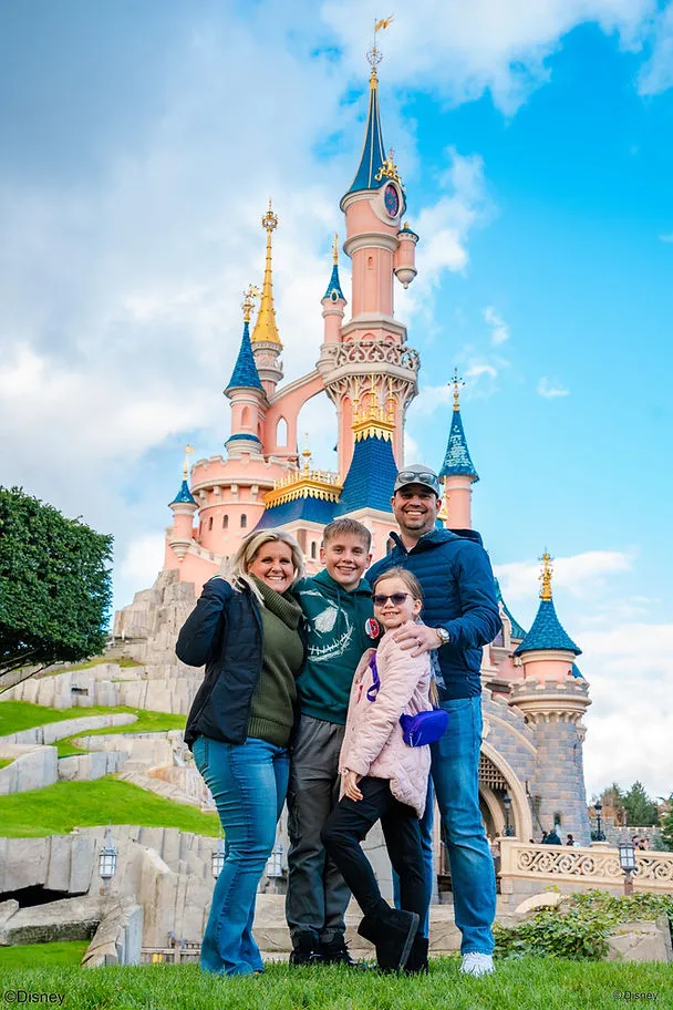 A family of four smiles in front of a pink and blue castle at Disneyland Paris. The sky is blue with clouds, and greenery surrounds the scene.
