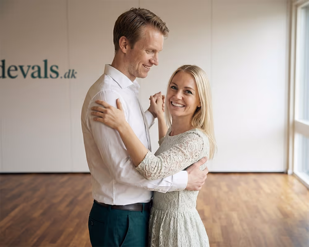 Smiling couple dancing indoors on wooden floor, woman in lace dress and man in white shirt.
