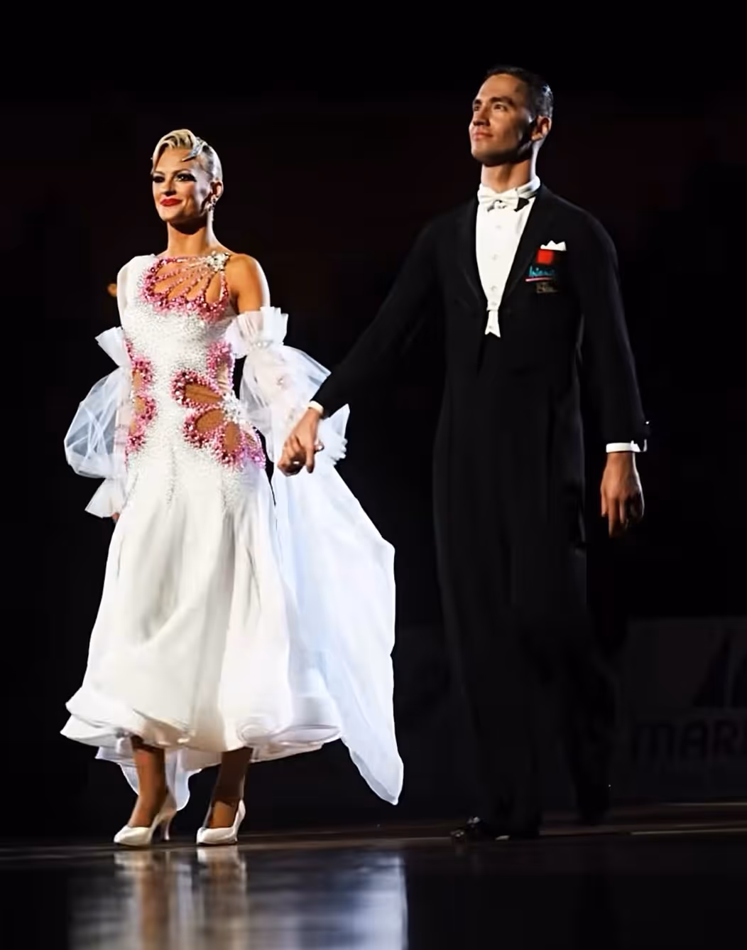 Signe Busk & Dmitri Kolobov. A ballroom dance couple holding hands on stage, the woman in a white dress with pink and white detailing and the man in a black tuxedo with a white bow tie.