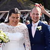 Smiling bride in a lace wedding dress holding a bouquet stands beside a groom in a navy suit with a white boutonniere, outdoors near a gazebo.