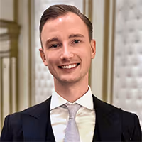 Oliver Curting. Smiling young man with short brown hair wearing a black suit, white shirt, and silver tie in an elegant room.