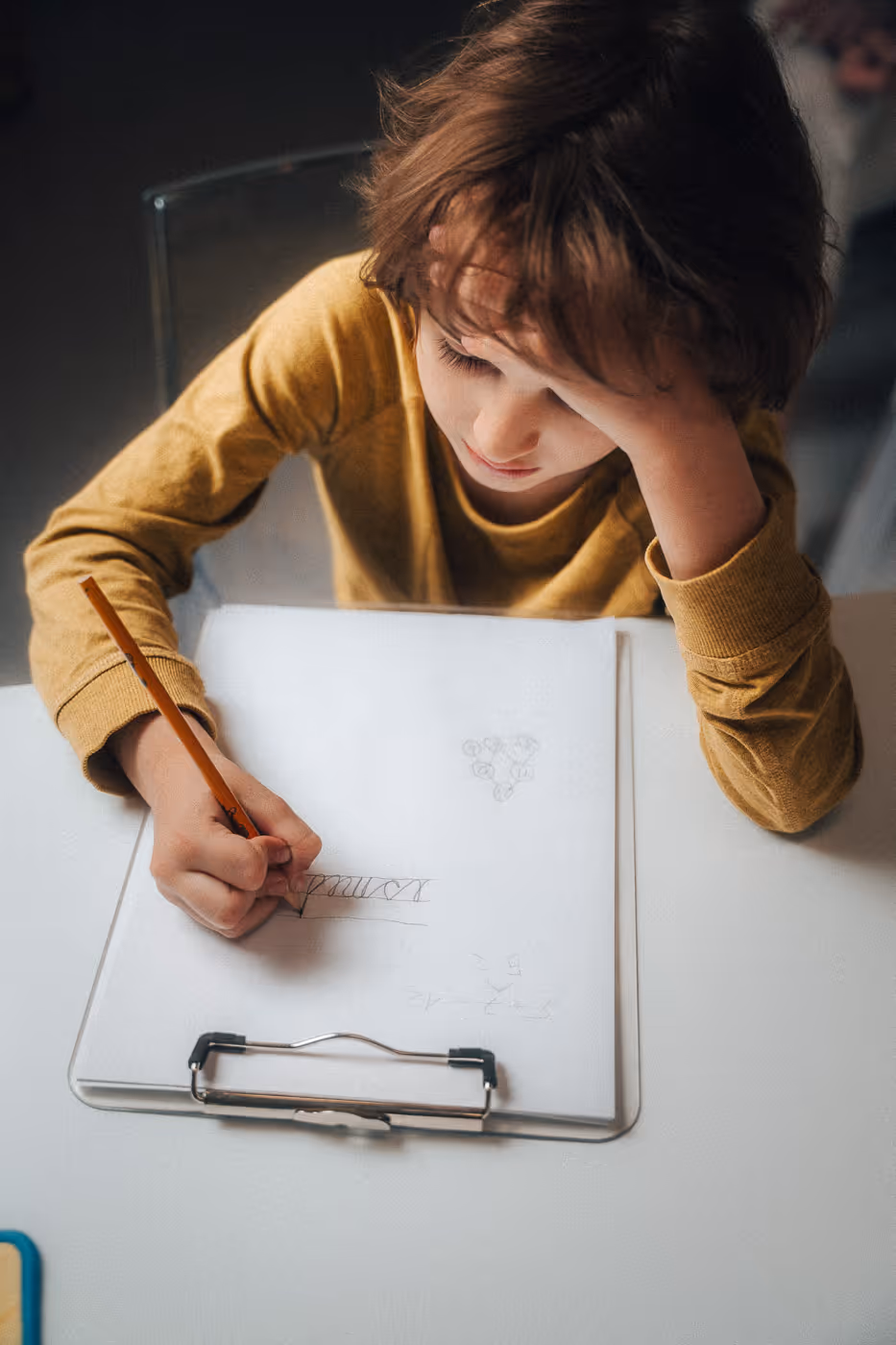 Young child in a mustard-yellow shirt writing on a sheet of paper clipped to a clipboard.