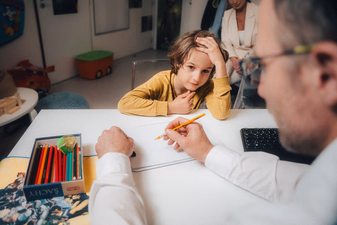 Child in a yellow shirt sitting at a table, looking at an adult holding a pencil and drawing on a tablet.