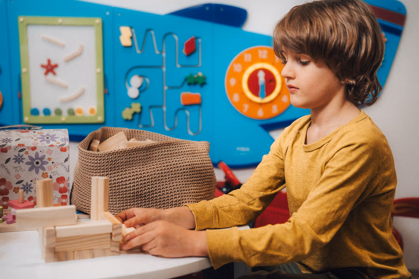 Young boy in a yellow long-sleeve shirt playing with wooden blocks at a white table in a colorful playroom.
