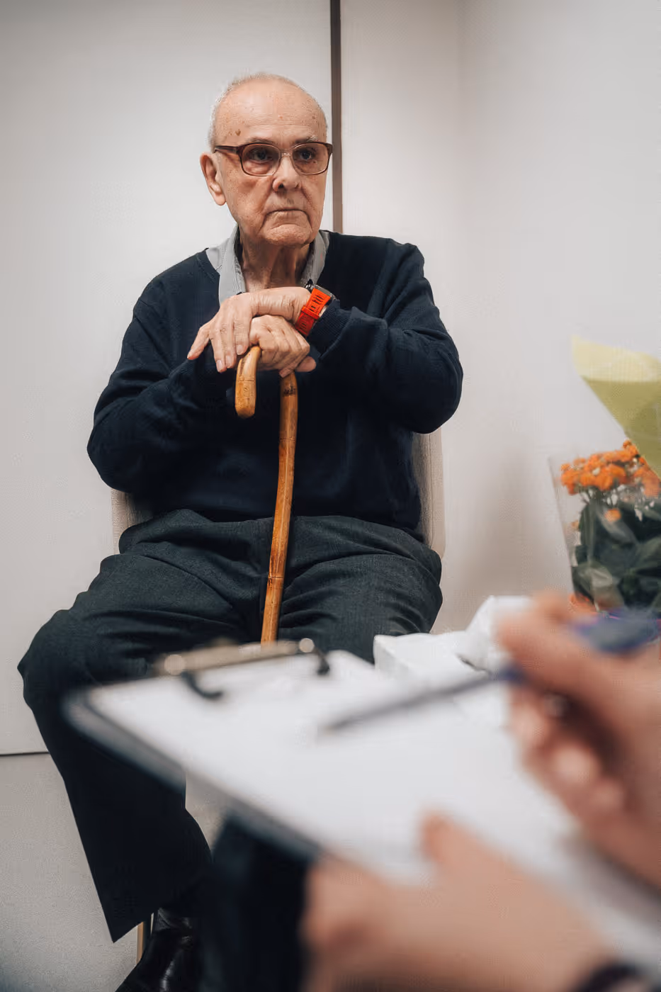 Elderly man with glasses sitting indoors holding a wooden cane, with a hand holding a pen and clipboard in the foreground.