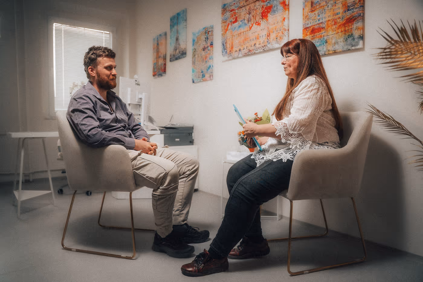 Man and woman seated facing each other in a brightly lit office with colorful paintings on the wall, engaged in conversation.
