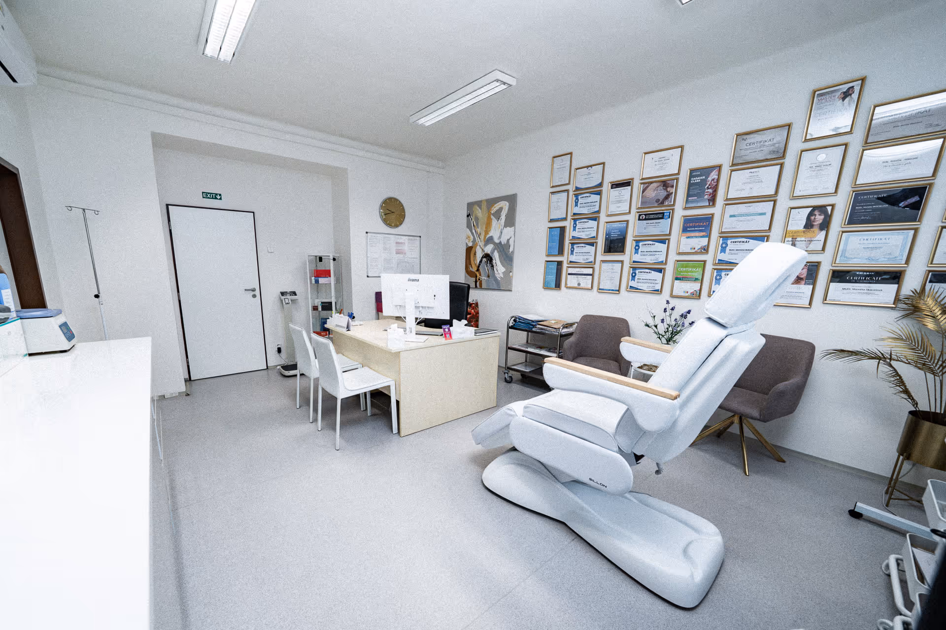 Bright and clean medical consultation room with a white examination chair, desk with computer, chairs, and wall displaying numerous framed certificates.