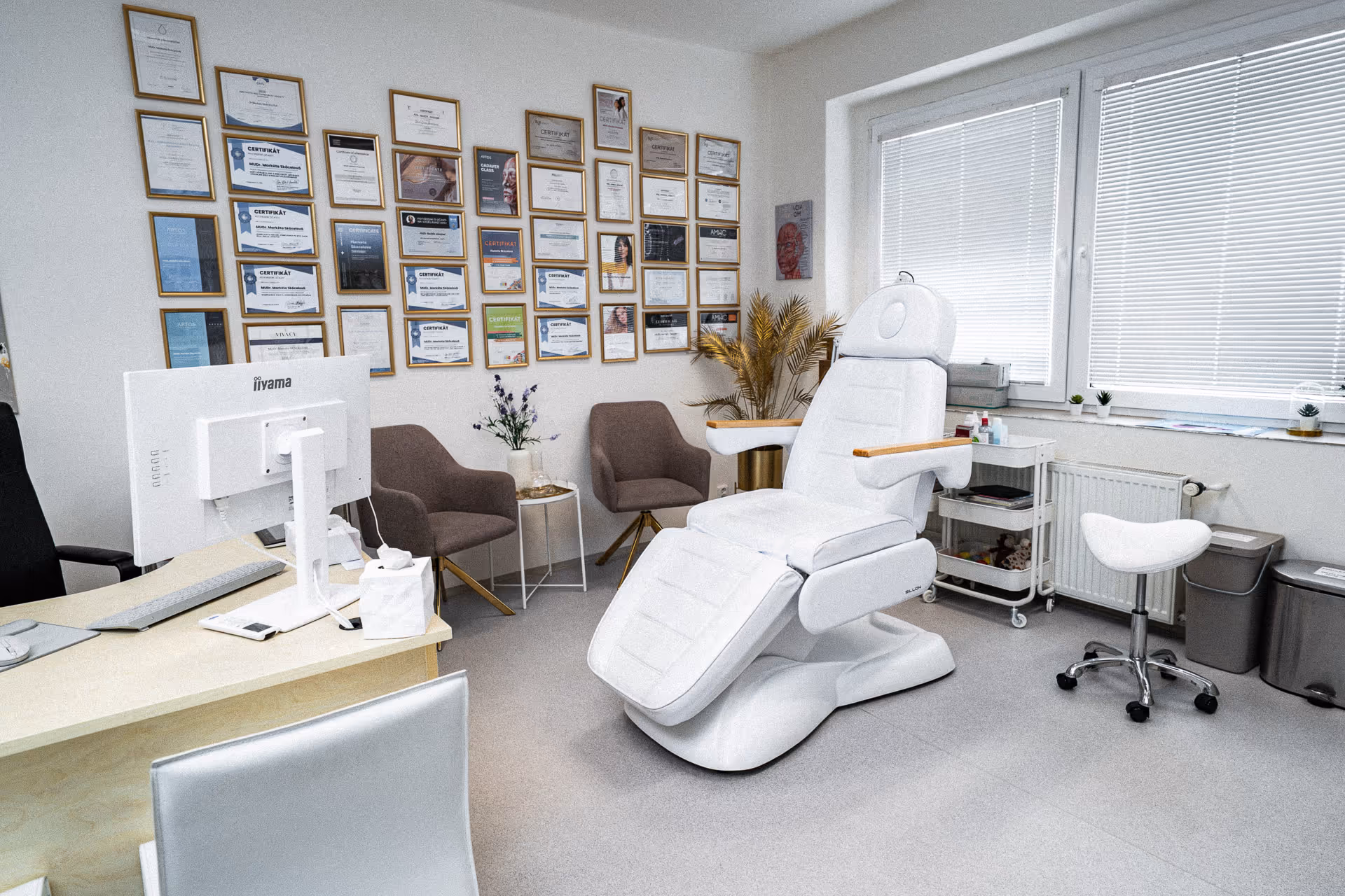 Modern medical consultation room with white examination chair, desk with computer, two brown chairs, and wall covered with certificates.
