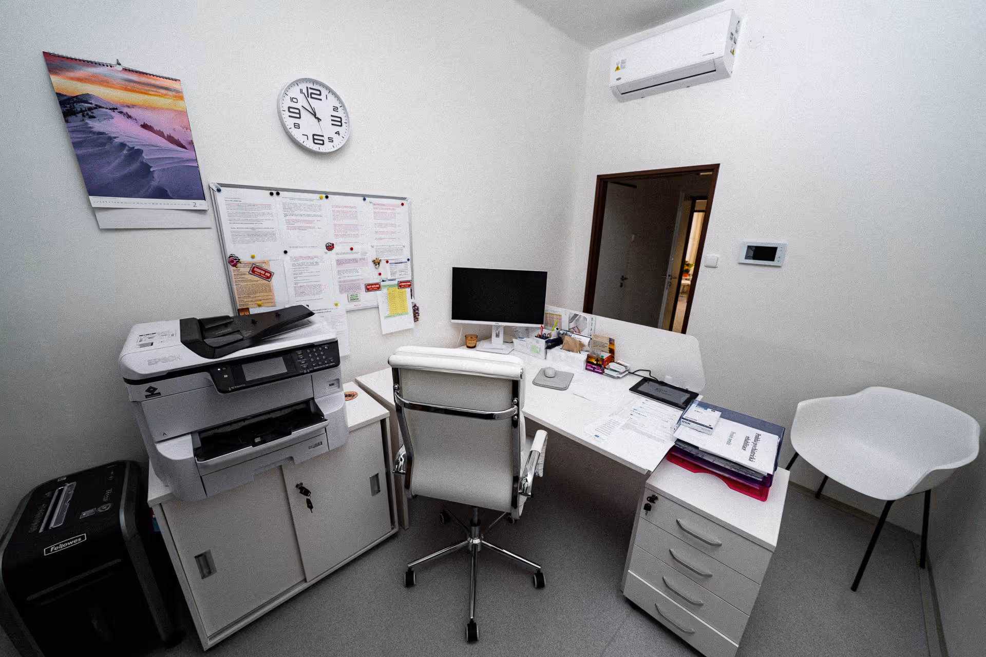 Small office corner with white desk, office chair, computer monitor, printer, calendar, wall clock, and documents.