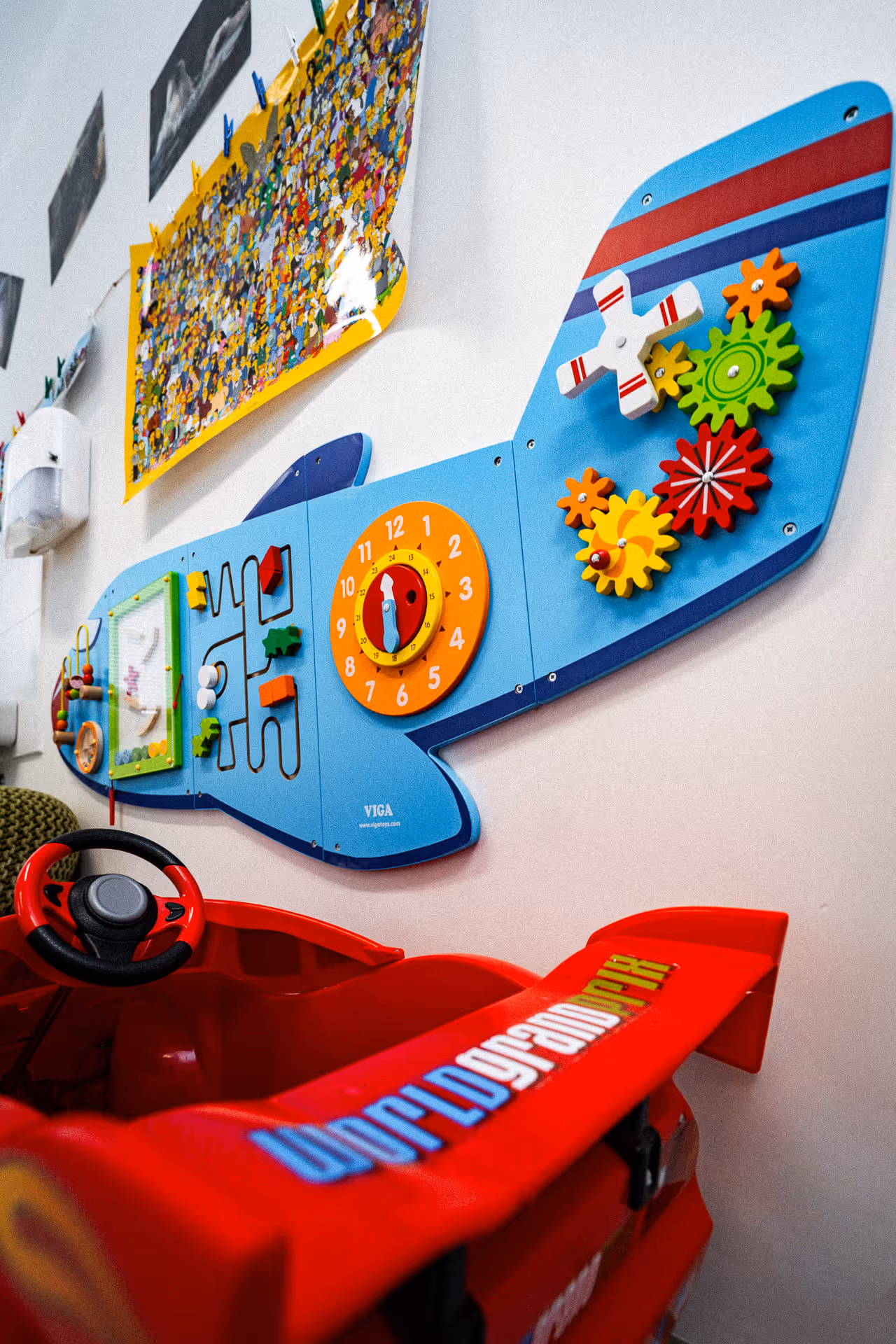 Child's play area featuring a red toy car and a blue airplane-shaped wall activity board with colorful gears, clock, and bead maze.