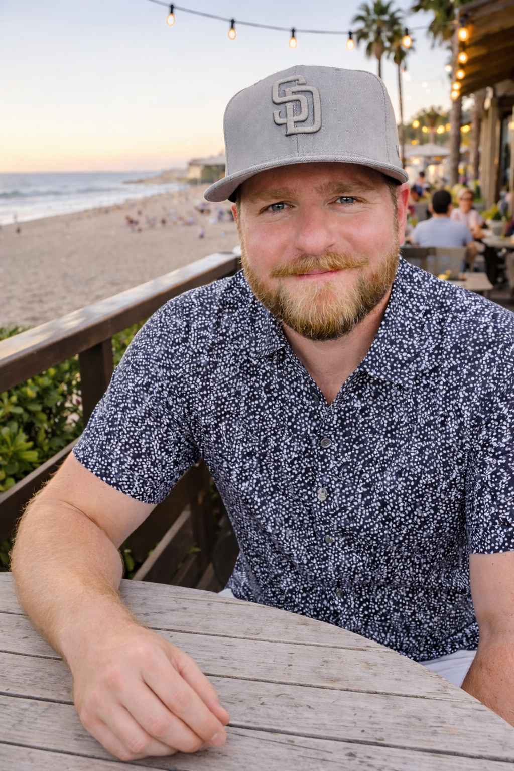 Man with a beard wearing a gray San Diego baseball cap and patterned shirt sitting at a wooden table by the beach at sunset.