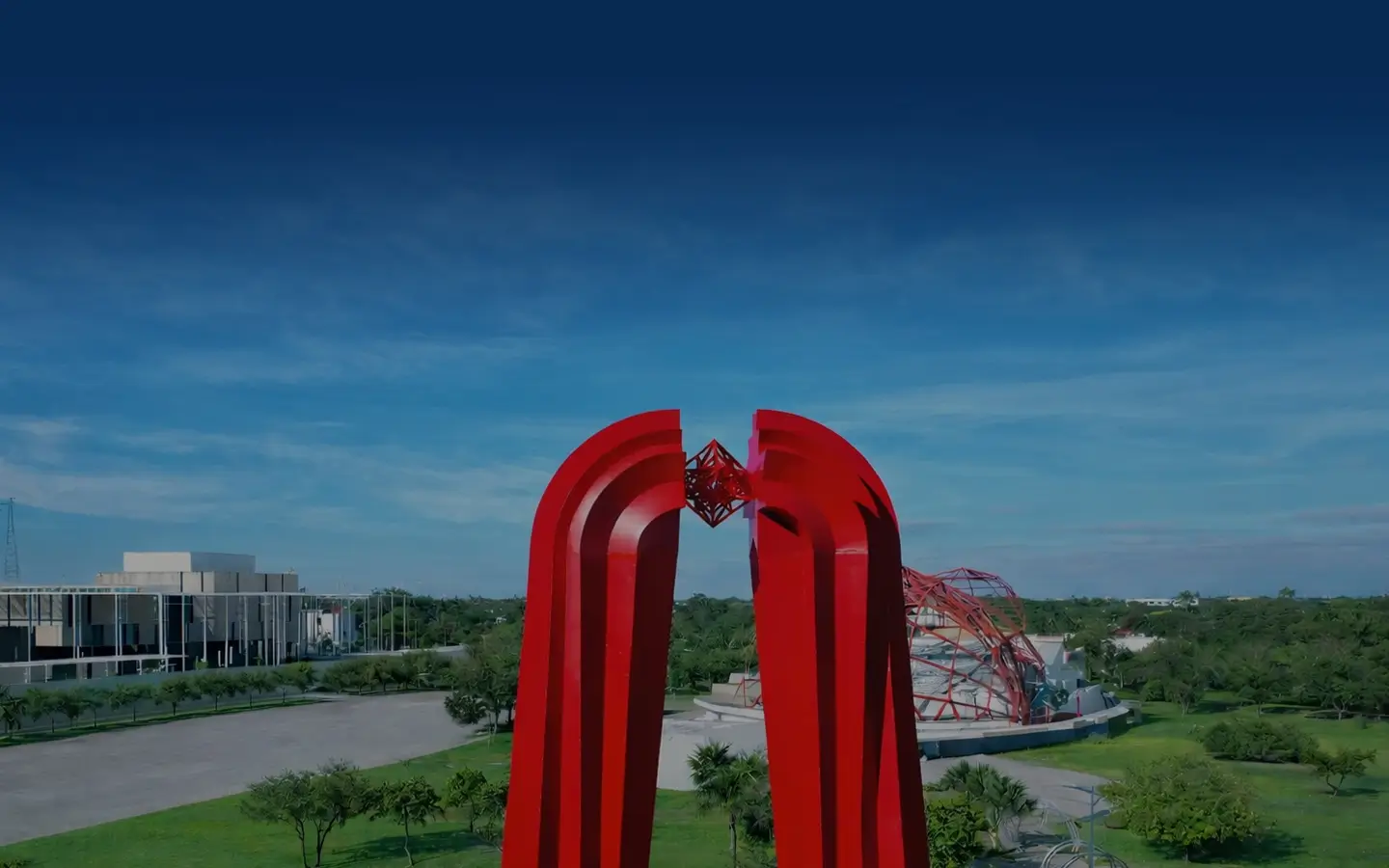 Red modern sculpture with a geometric centerpiece in a green park under a blue sky.