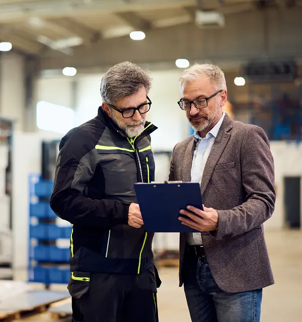 Two middle-aged men wearing glasses reviewing documents on a clipboard in an industrial warehouse.