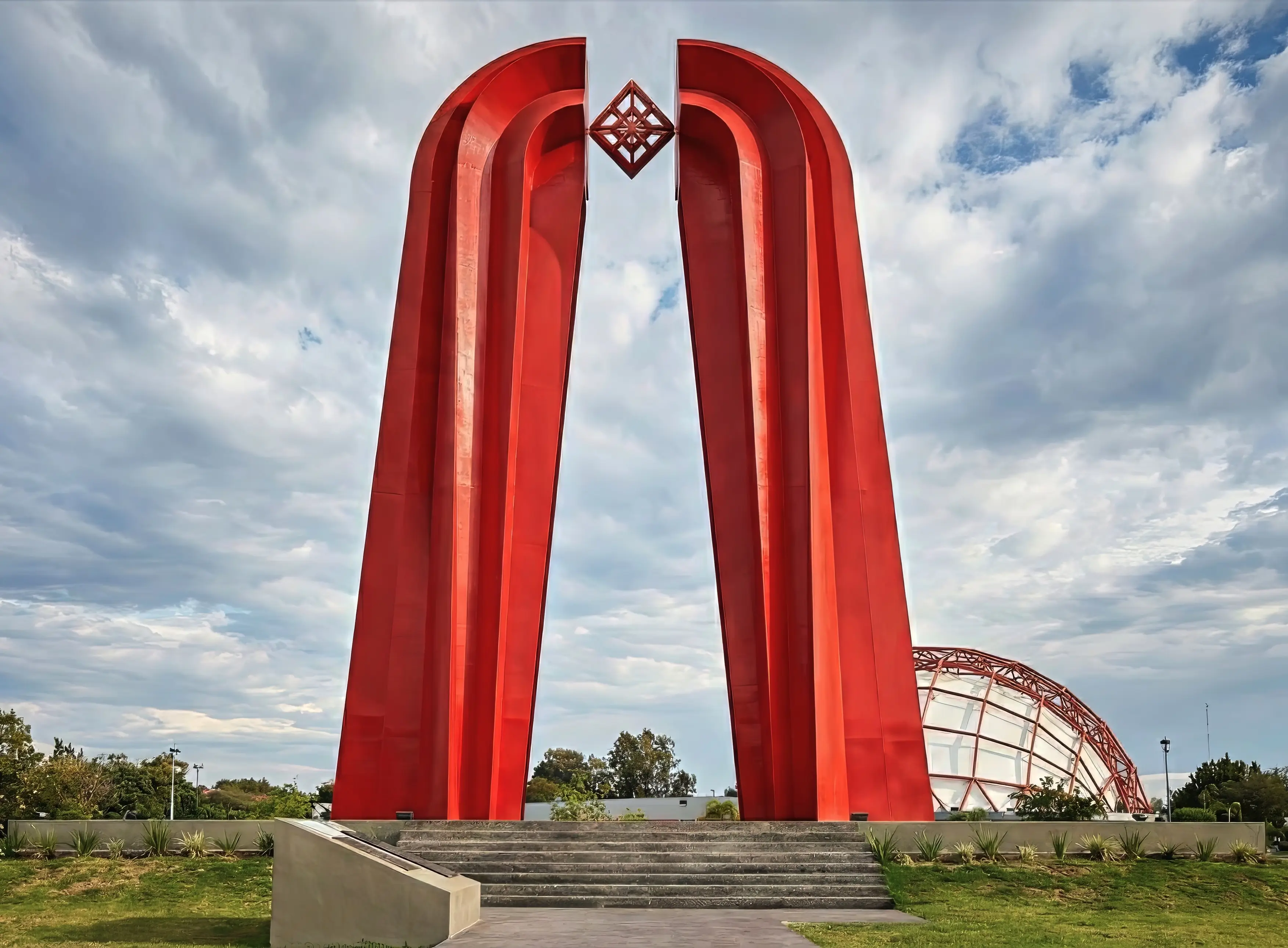 Tall red metal arch sculpture with a geometric diamond shape suspended at the top, set against a partly cloudy sky and green grass.