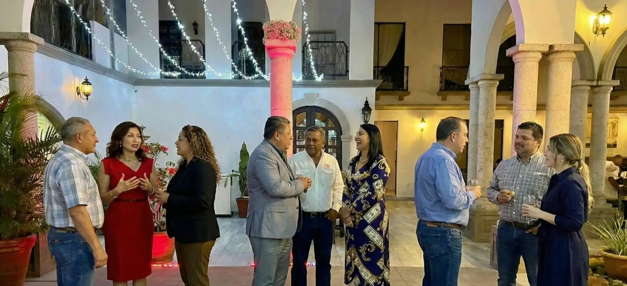 Eight people in business casual attire engaged in conversation in a warmly lit courtyard with arches and string lights overhead.