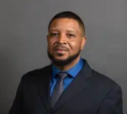 A headshot of a black man wearing a a blue suit and tie