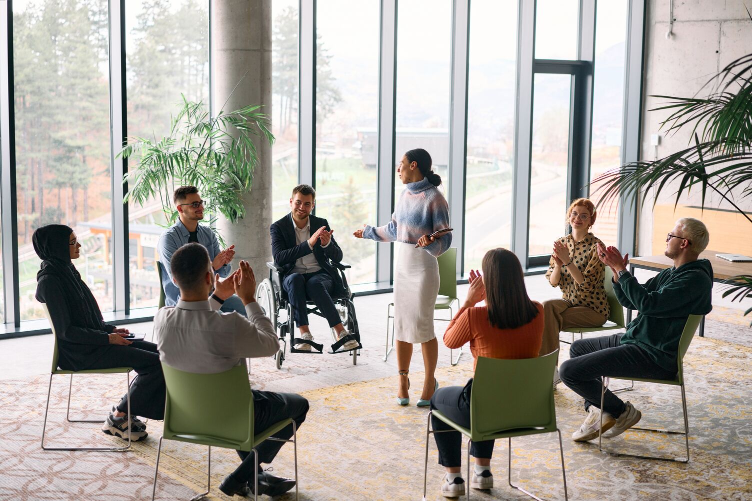 Diverse group of adults seated in a circle applauding a standing woman speaking in a bright room with large windows and plants.