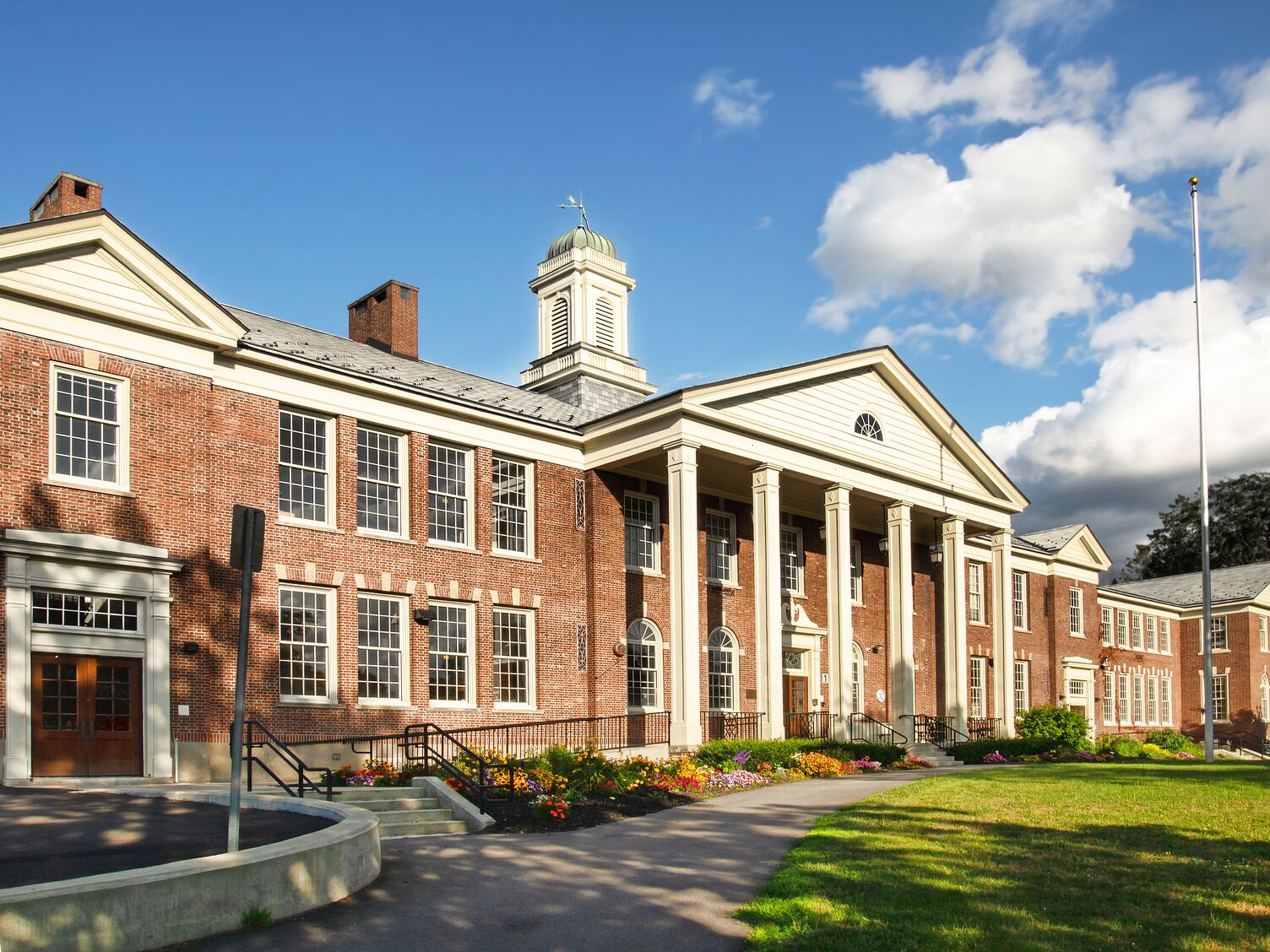 Classic American school building with red brick walls, white columns, a cupola, and landscaped flower beds under a blue sky with clouds.