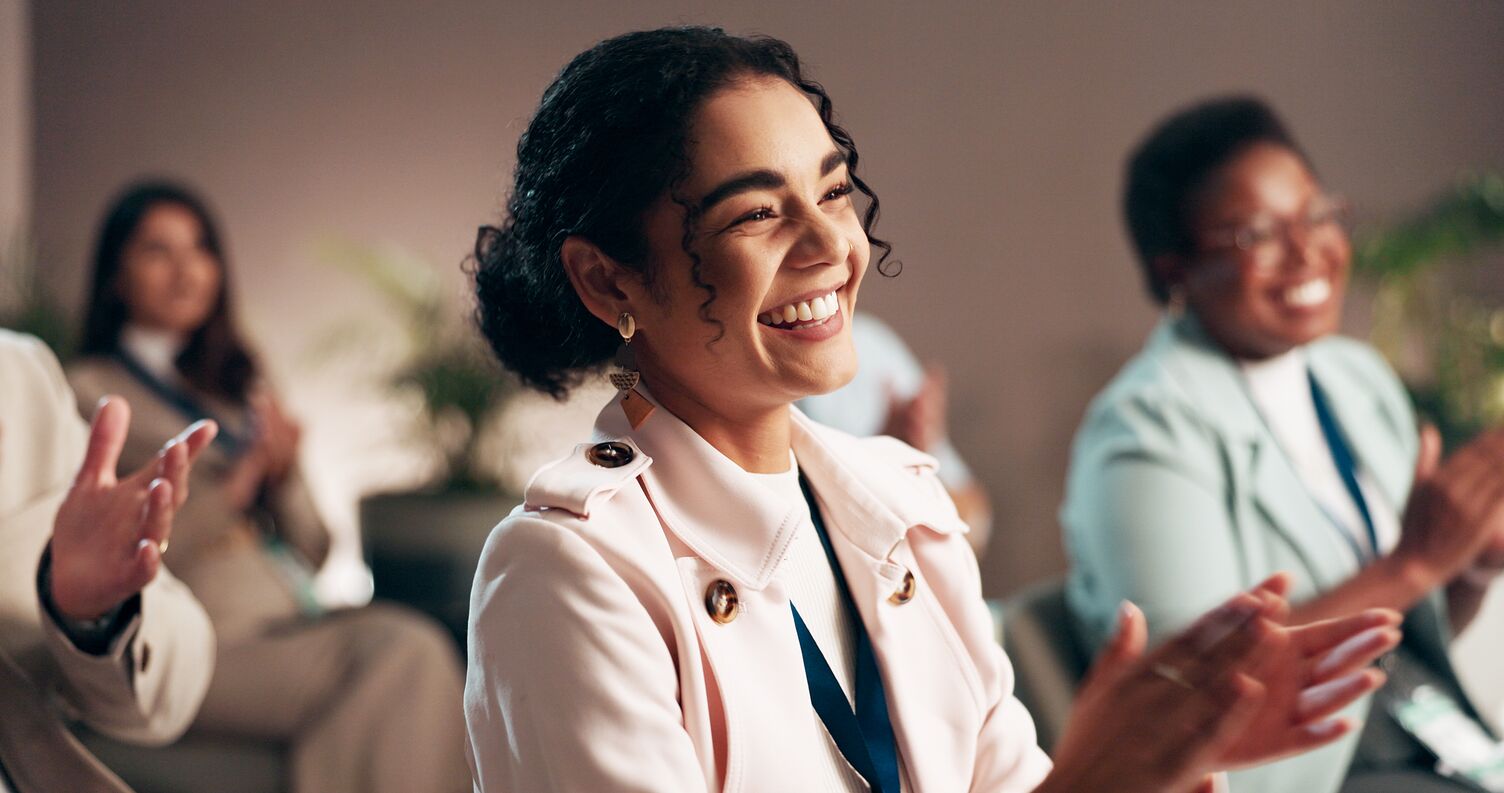 Smiling woman in light-colored coat applauding during an indoor event with others clapping in the background.