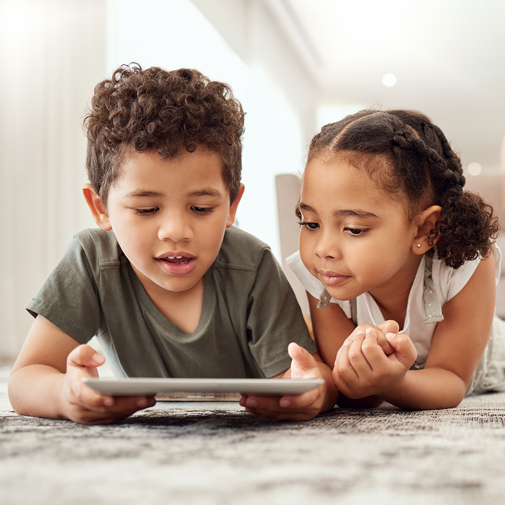 Two children lying on a carpet together, looking at a tablet screen.