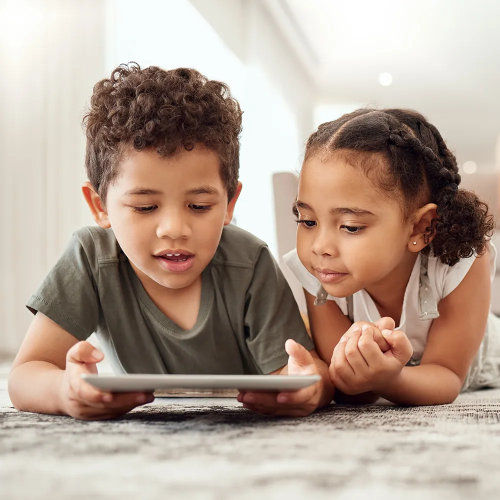 Two children lying on a carpet together, looking at a tablet screen.