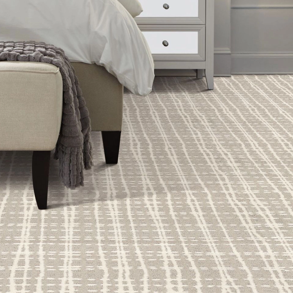 Beige carpet with white linear pattern under a beige bed frame with a dark gray textured blanket and a gray nightstand with white drawers.