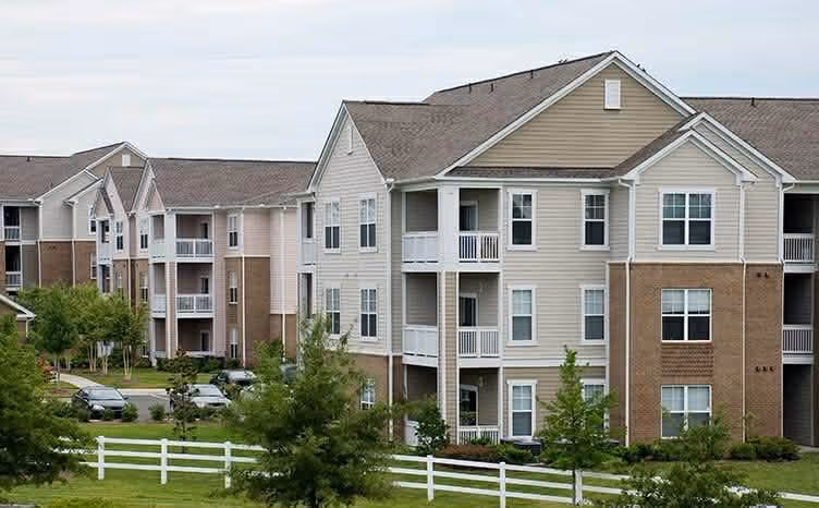 Three-story residential apartment buildings with beige and brown siding, balconies, and a white wooden fence with green trees and grass in the foreground.