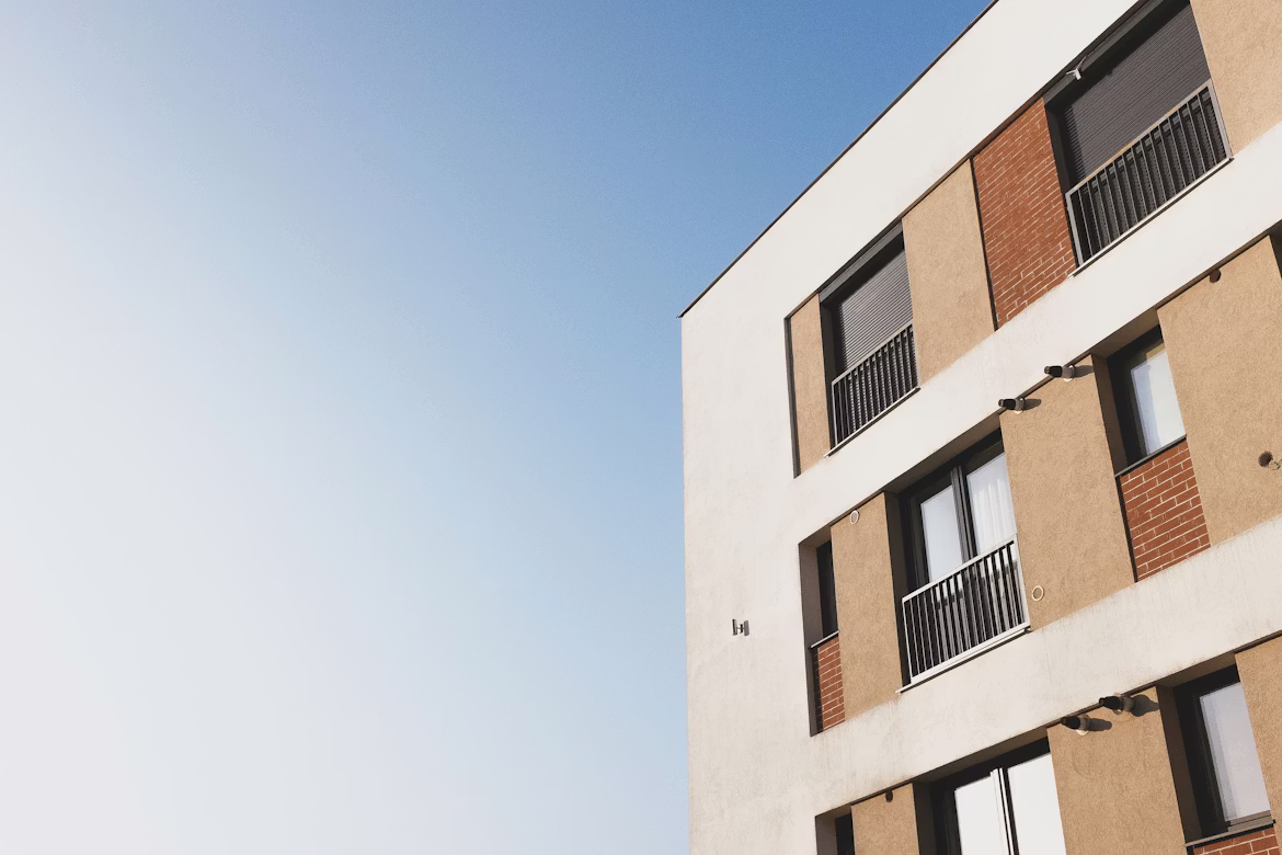 Modern building facade with beige and brick sections featuring windows and balconies against a clear blue sky.