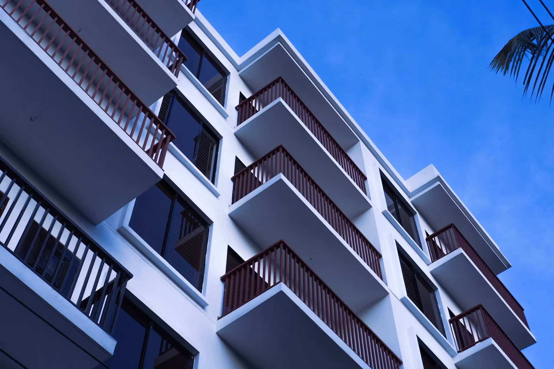Low-angle view of a modern white apartment building with multiple balconies and dark railings under a clear blue sky.