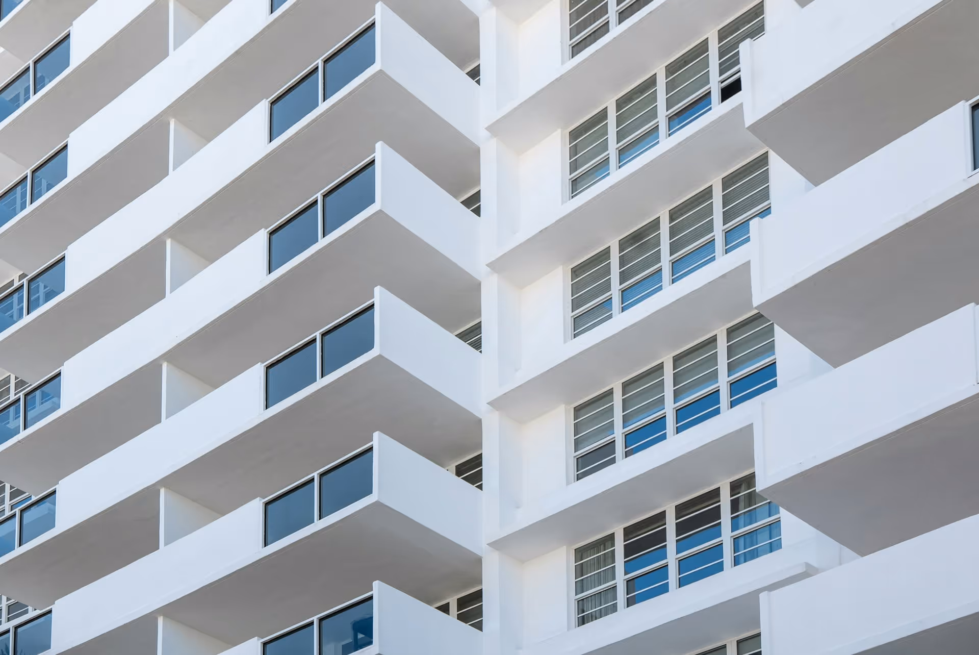 Modern white building facade with rows of balconies and reflective windows under clear sky.