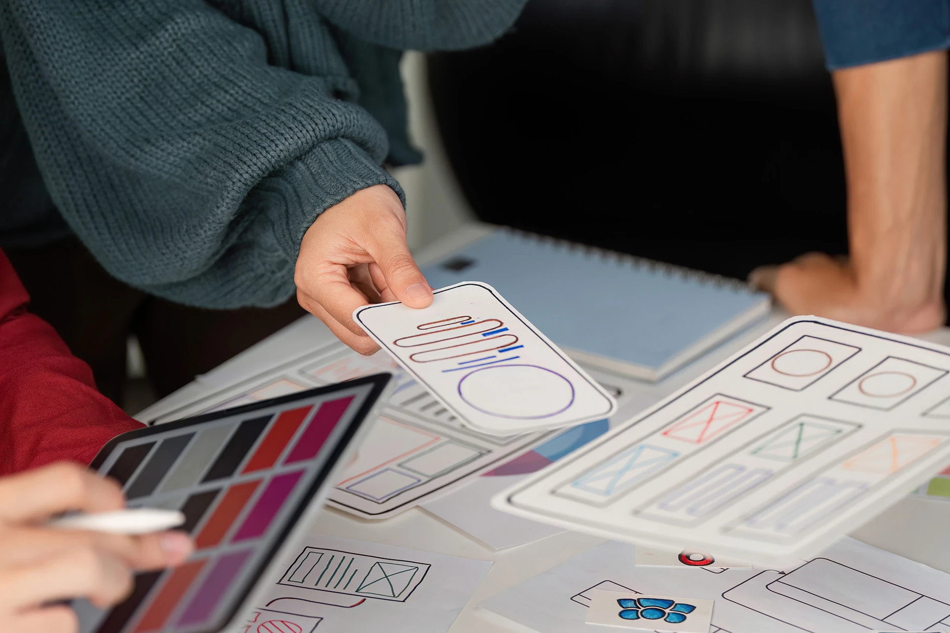 Close-up of hands arranging paper UI wireframe cutouts and color palette swatches on a table during a design session.