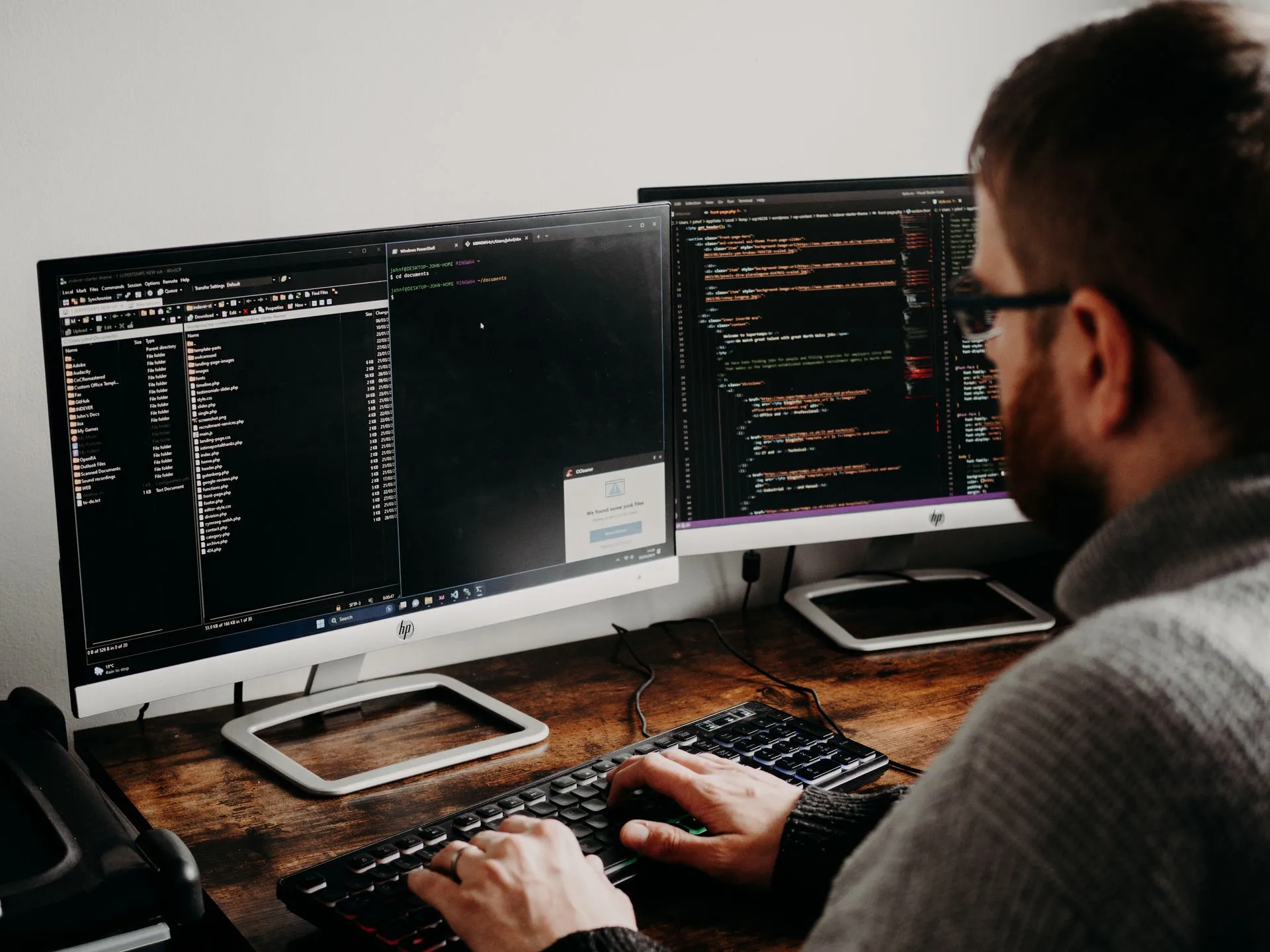 Developer typing on a keyboard while reviewing code across two monitors at a desk.