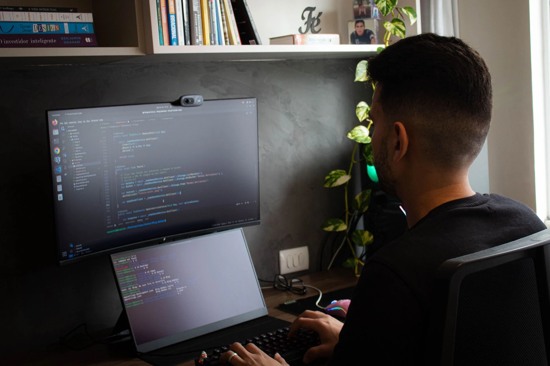 Developer coding at a dual-monitor desk setup in a dim home office with plants and a bookshelf.