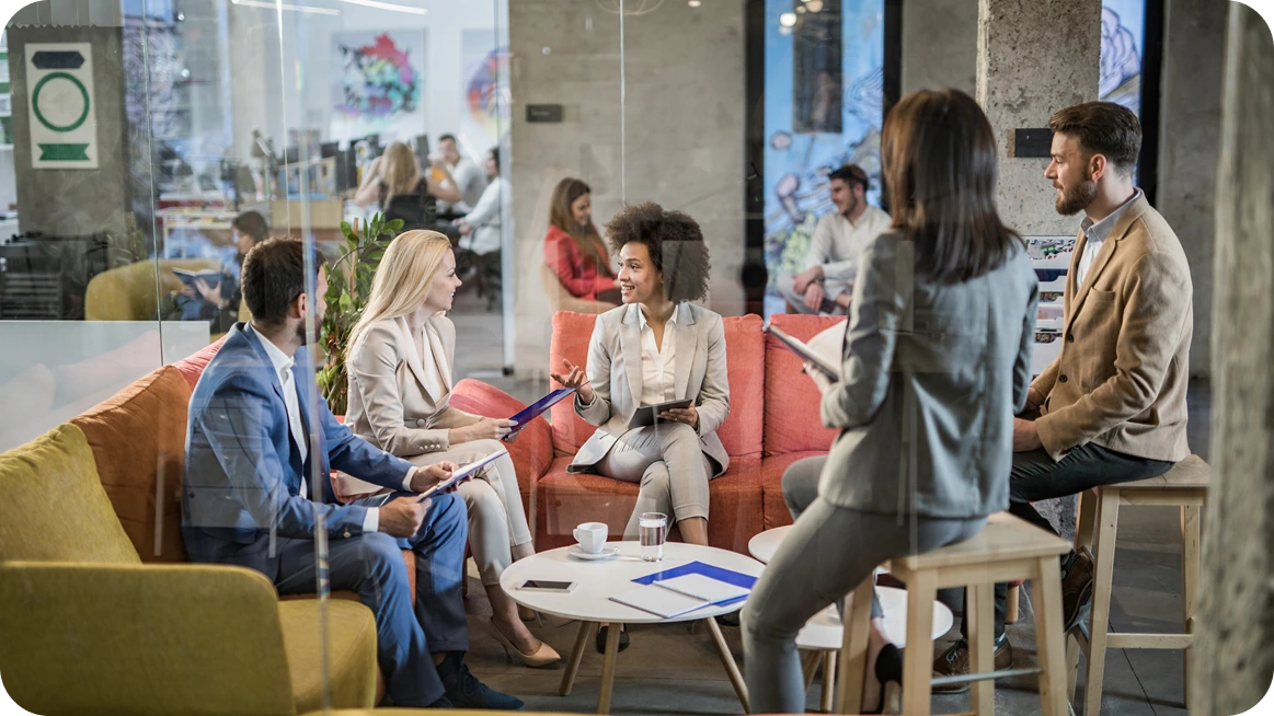 Five diverse colleagues in business attire engaged in a meeting around a coffee table in a modern office lounge.