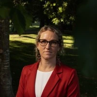 Woman with glasses wearing a red blazer and white top, standing outdoors with greenery in the background.