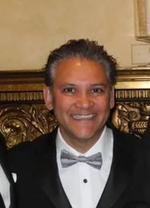 Smiling man in a black tuxedo and gray bow tie with ornate wooden background.