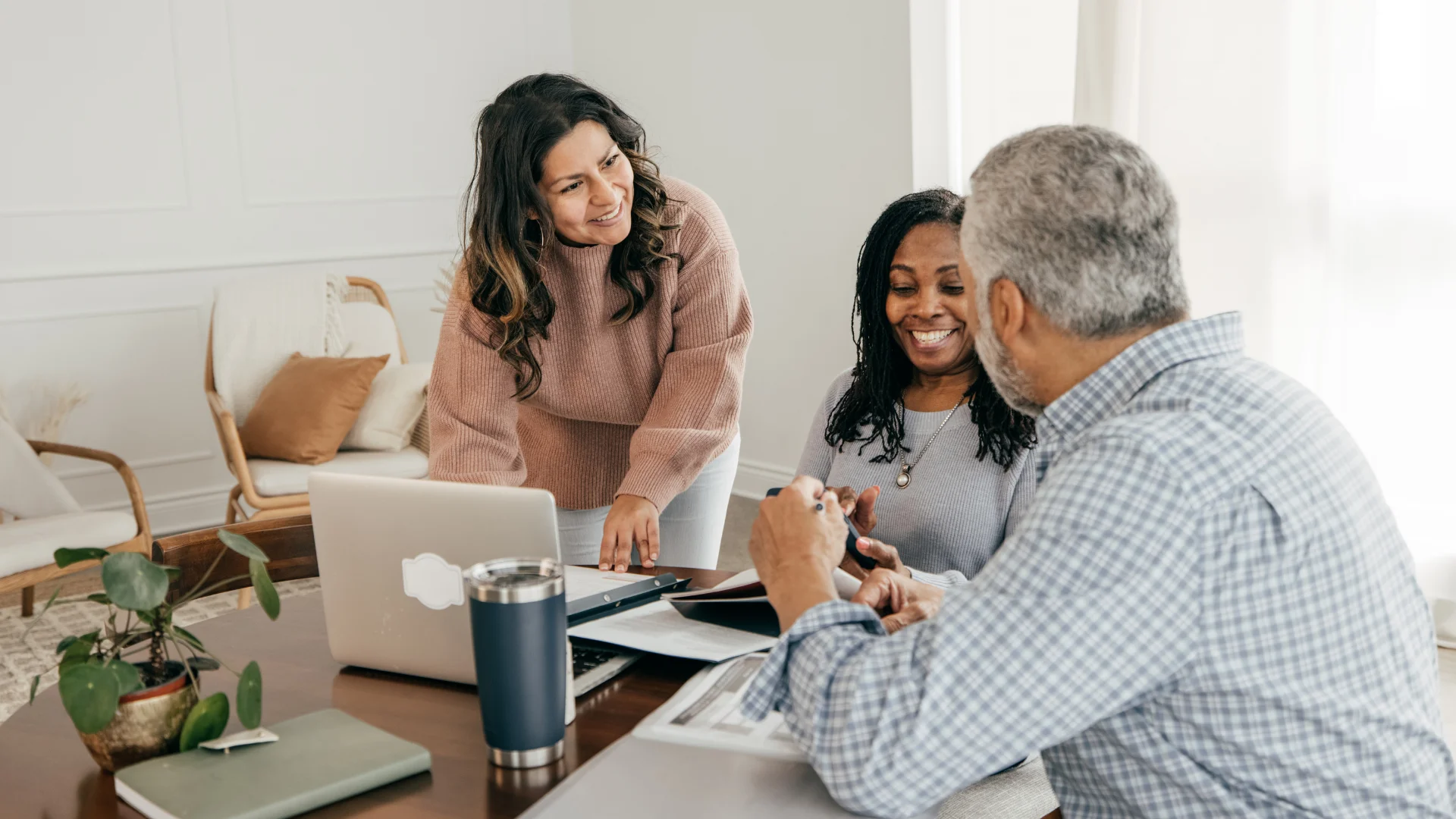 Three diverse people smiling and discussing documents at a table with a laptop and plants in a bright room.