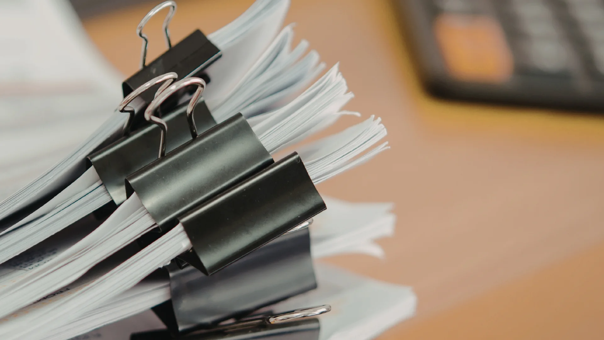 Stacks of white papers held together with black binder clips on a wooden desk with a blurred calculator in the background.