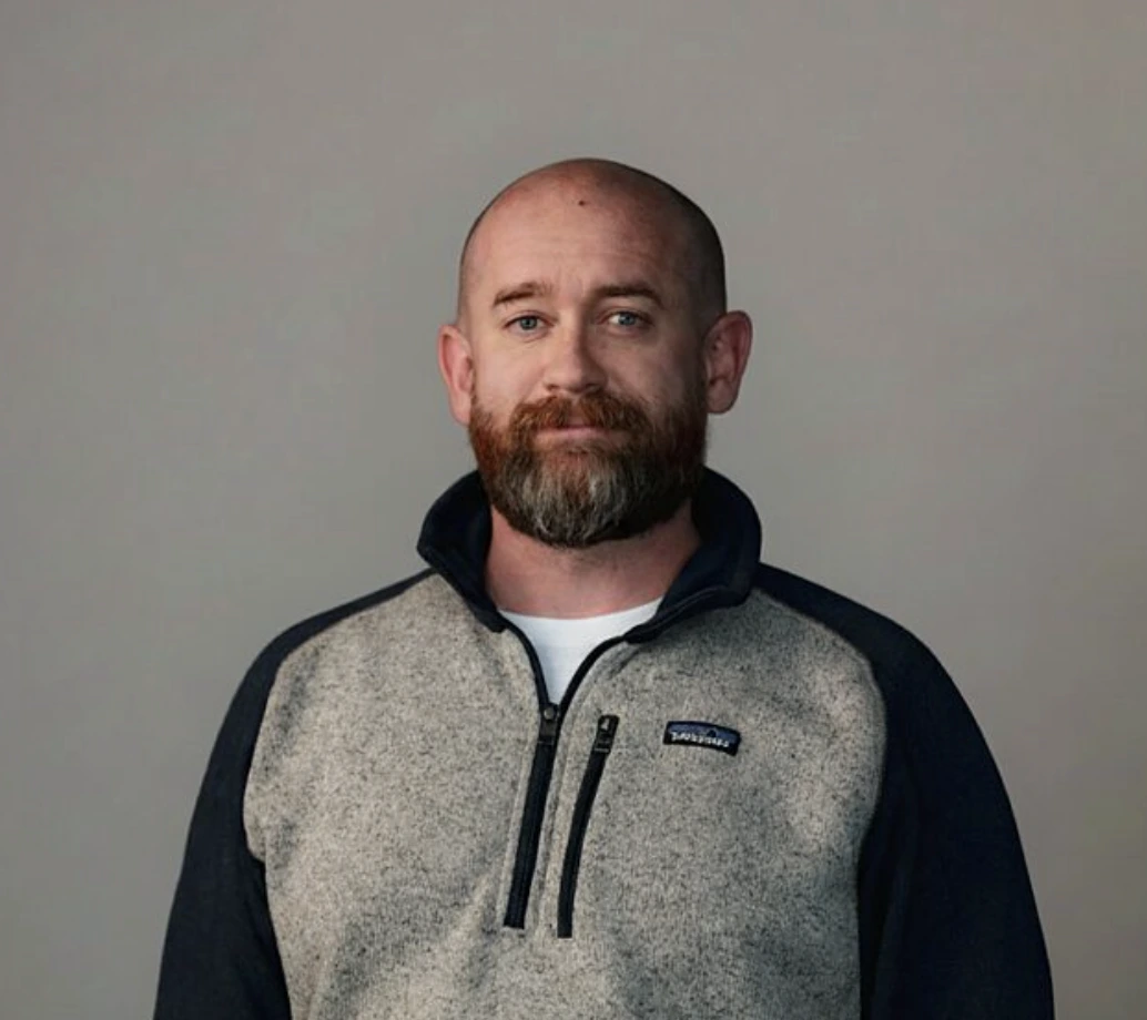 Portrait of a bald man with a beard wearing a gray and black zip-up sweater against a neutral background.