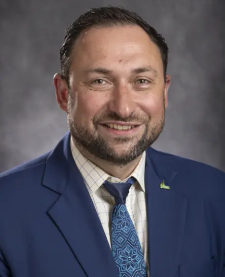Smiling man with short dark hair and beard wearing a blue suit jacket, patterned blue tie, and a checkered shirt against a gray background.