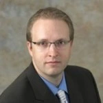 Man with short light brown hair, wearing glasses, a black suit, light blue shirt, and striped tie, posing against a grayish background.