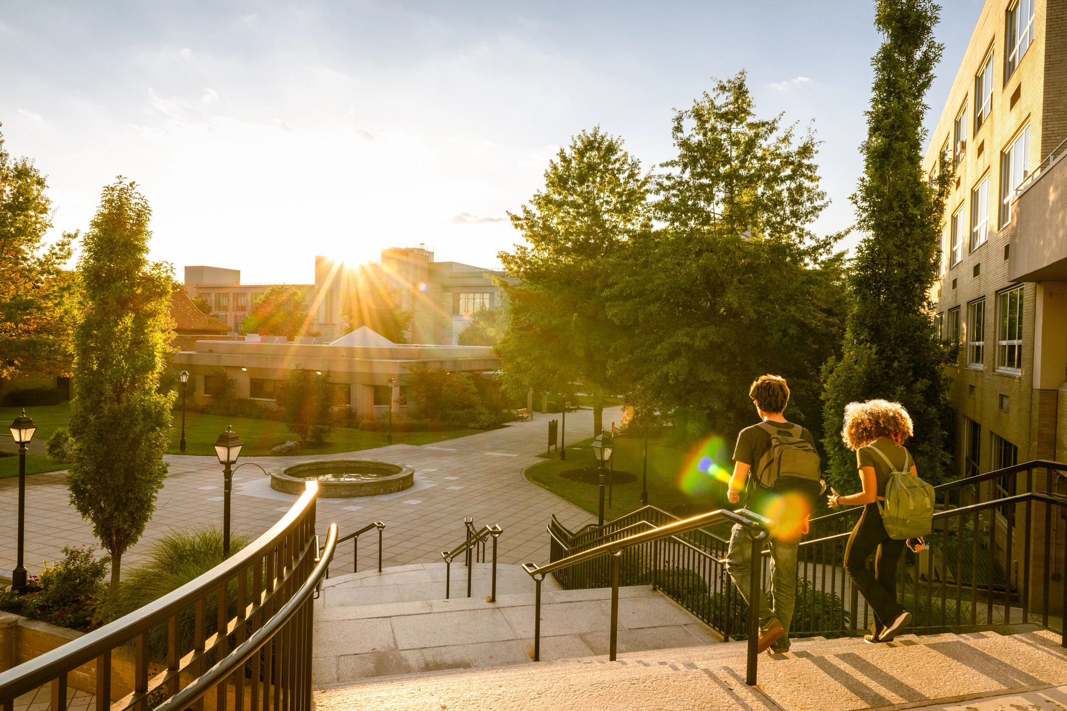 Two students with backpacks walking down stairs on a university campus at sunset with trees and buildings in the background.