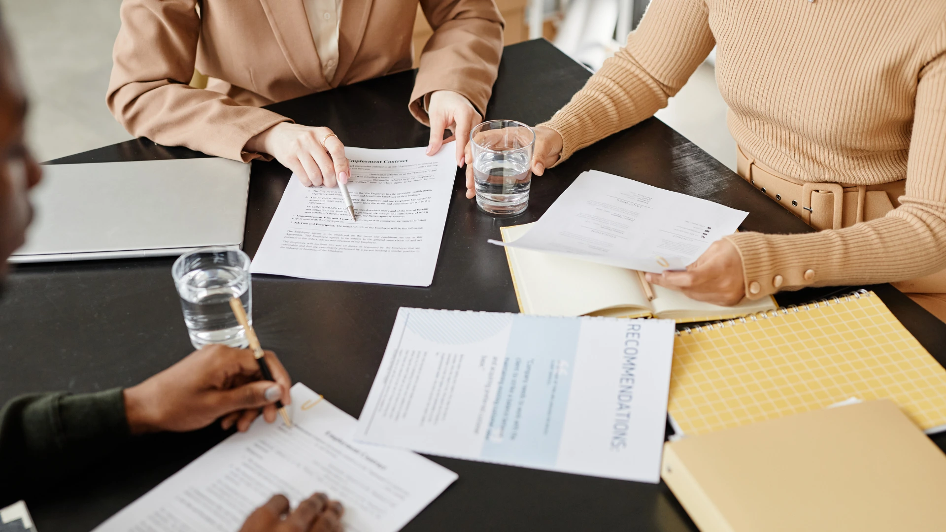 Three people sitting at a table reviewing documents, including an employment contract and recommendations letter, with glasses of water and a laptop.