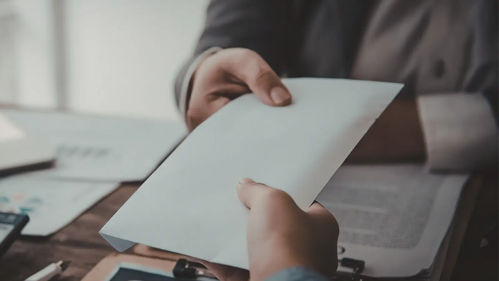 Two people exchanging a white envelope over a desk with documents and a calculator.