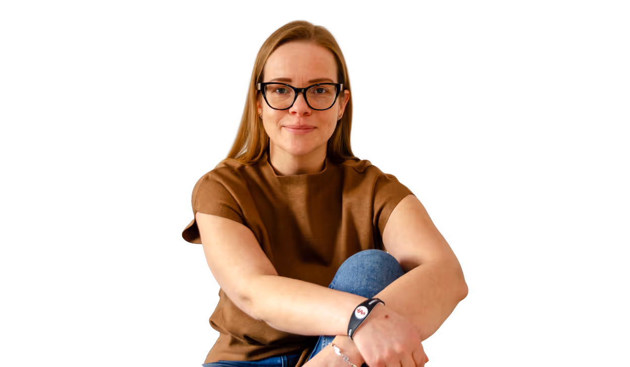 Frau mit braunem Oberteil, Jeans und Brille sitzt mit verschränkten Armen und lächelt leicht.