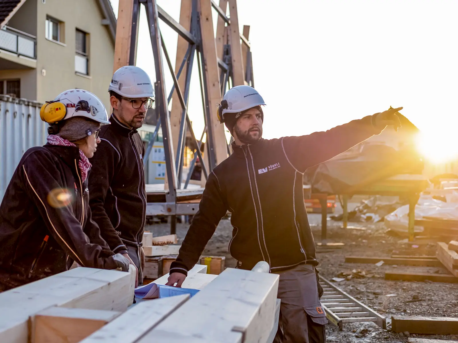 Drei Bauleiter auf Holzbaustelle bei Sonnenuntergang, Besprechung und Koordination des Montageablaufs mit präziser Planung.