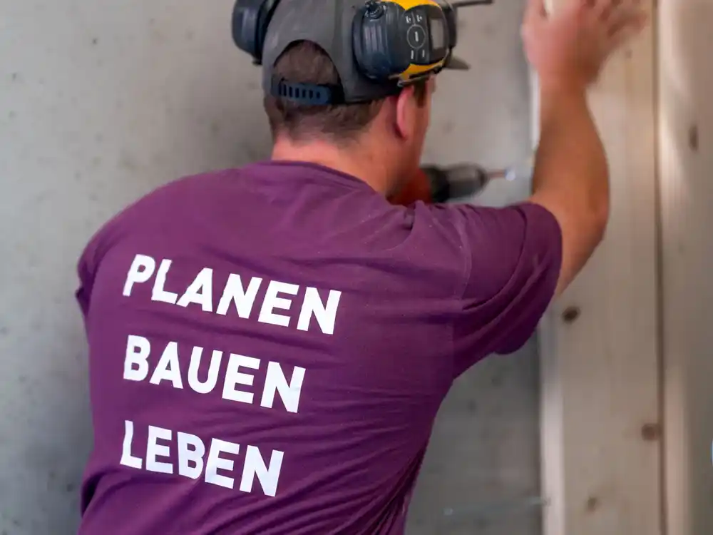Handwerker mit gelbem Helm und Ohrenschützern bohrt in einer Wand, trägt T-Shirt mit Text Planen Bauen Leben.